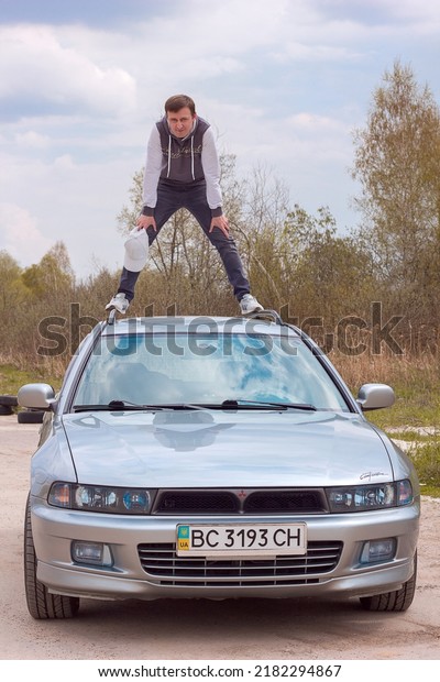 Lviv Ukraine - 05 02 2022: A young man in stylish\
clothes and a cap stands on the roof of a Mitsubishi Galant car\
against the background of the road and trees, beautiful and fast\
car speed and comfort