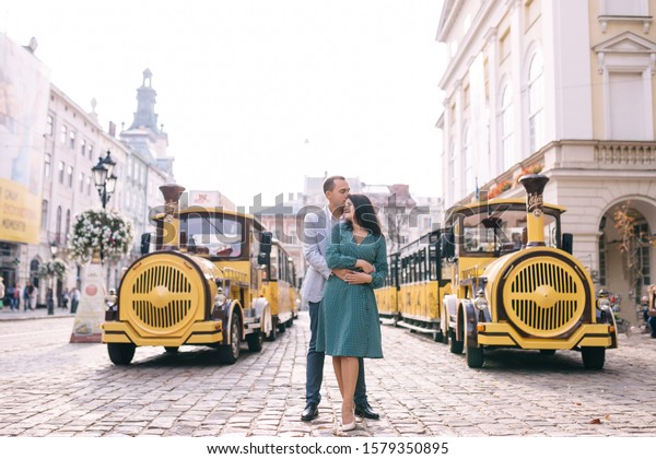 Lviv, Ukraine - 23 September, 2019 husband hugs\
wife from the back side and they look at each other. locomotive and\
wagons on the city\
square.