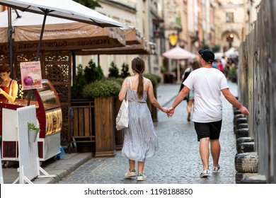 Lviv, Ukraine - July 30, 2018: Back Of Young Romantic Couple Holding Hands Walking In Historic Ukrainian Polish Lvov City During Evening, Outdoor Cafe Ice Cream Restaurant, Street In Old Town