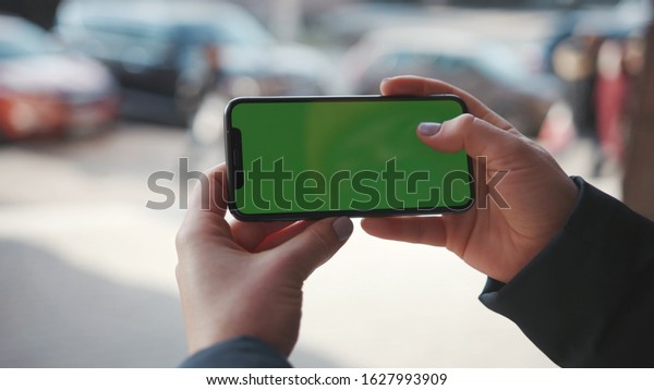 Lviv, Ukraine - May 19, 2018: Close up shot of woman\
hands holding phone with horizontal green screen on the city street\
background sunset people car busy finger touch message cellphone\
display girl