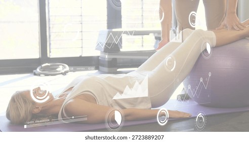 Lying client stabilizing legs on purple mat and ball in studio with HUD, wearing tanktop, leggings. Trainer, trainee, fitness, gym, balance, core, daylight