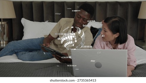 Lying couple using tablet and silver laptop on bed, wearing beige tee, pink top, holographic data. Augmented, reality, bedroom, modern, minimalist, collaboration, techsavvy