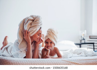 Lying down on white bed together. Young mother with her daugher have beauty day indoors in room.