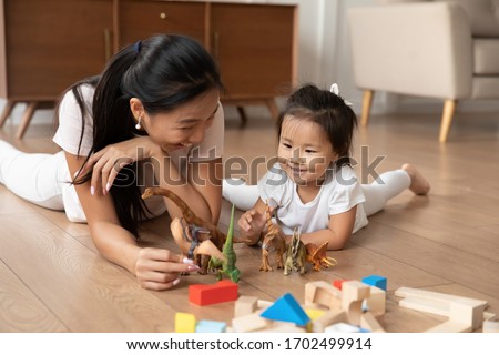 Lying on warm wooden floor asian mom play game with little daughter holds dinosaurs toys heap of colorful construction blocks nearby. Spend time have fun together educational activity with kid concept