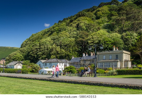 Lynmouth, Devon, England, 13 July 2016: People\
walk along the path past the small houses, otlele and cafes. Two\
sitting on the bench. Sea\
shore.