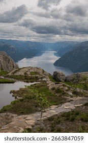 Lysefjord extends between steep cliffs near Preikestolen, Norway. Rugged terrain, greenery, and a small pool of water appear under a cloudy sky.