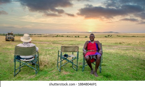 Maasai Mara National Reserve, Kenya - September 29, 2013. A Kenyan Safari Guide And An American Male Guest Relax On A Game Drive Rest Break, As The Sun Sets Over The Vast African Landscape.