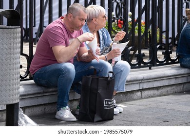 MAASTRICHT, THE NETHERLANDS - MAY 28 2021: Slightly Overweight Man And His Wife Sit In A Street On A Wall In Front Of A Fence In The Sun And Eat Chips From A Paper Cone