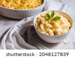 Mac and cheese in white bowl with basil on top and another mac and cheese baked in oven in background placed on a white rustic board