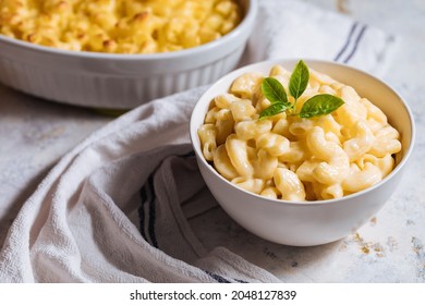 Mac and cheese in white bowl with basil on top and another mac and cheese baked in oven in background placed on a white rustic board