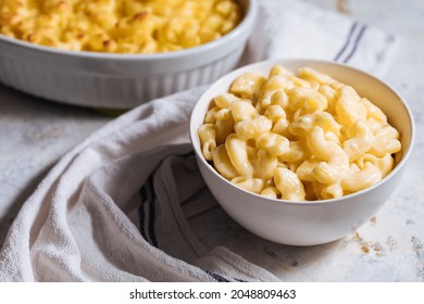 Mac And Cheese In White Bowl With Basil On Top And Another Mac And Cheese Baked In Oven In Background Placed On A White Rustic Board