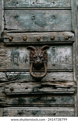 Macabre and unusual door knocker on a faded weathered door. The head of a lion with goat horns depicts a chimera and looks creepy, but means strength, independence and stubbornness.