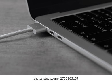 A MacBook is connected to a charging cable through its USB port. The device sits on a wooden table, indicating it is in use and charging.