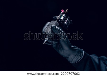 Machine for tattoo. Human hand in black protective glove holding professional equipment for making tattoo on body isolated on dark background. Concept of art, job, hobby, poster for ad