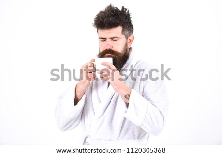 Macho drowsy, sleepy face drinks coffee in morning enjoying aroma. Man with beard and disheveled hair stands in bathrobe, holds mug with tea or coffee, white background. Morning rituals concept.