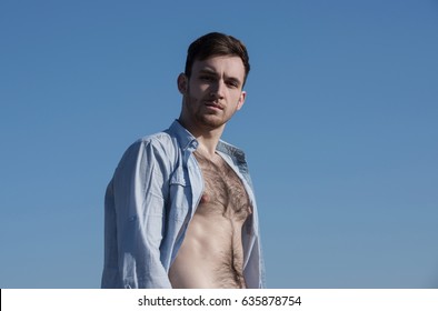 macho. Handsome, sexy, young, caucasian man or model with bearded face and stylish hair, haircut, posing in unbutton shirt with hairy, naked torso on sunny, summer day, on blue sky background.
