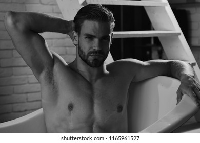 Macho sitting naked in bathtub with romantic light on, selective focus. Man with beard and serious face. Guy in bathroom with stairs on background. Sex and erotica concept. 
