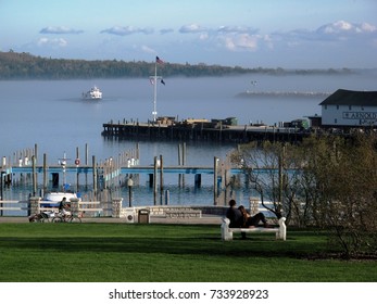 MACKINAC ISLAND, MI: May 20, 2011 - Young Couple Lounges On A Park Bench Watching The Ferries Come In To The Harbor On A Foggy Day
