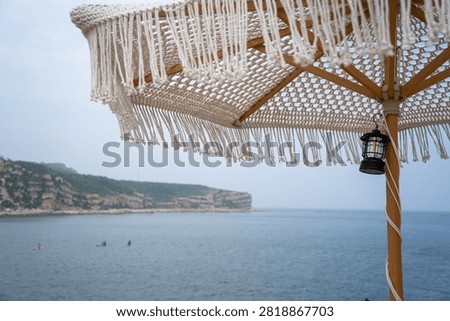 A macrame beach umbrella frames a tranquil seascape with a rocky cliff, calm water, and small watercraft under an overcast sky.