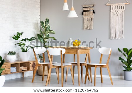 Macrame hanging on gray wall above wooden table and chairs in bright dining room interior with lots of plants. Real photo