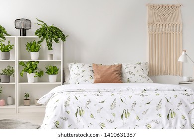 Macrame Hanging On The Wall In Real Photo Of White Bedroom Interior With Many Fresh Plants On Rack And Double Bed With Floral Sheets