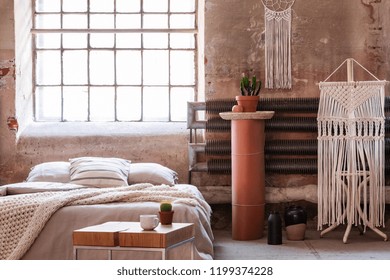 Macrame In A Wabi Sabi Bedroom Interior With A Bed, Table And Stand With A Plant. Real Photo