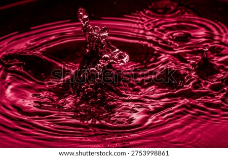 Macro capture of a dramatic water splash forming a rising droplet column with circular ripples across a vivid red liquid surface, highlighting dynamic motion and glossy reflections