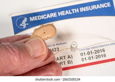Macro close up of fingers holding tiny modern hearing aid on medicare card background as healthcare coverage is improved - Shutterstock ID 2063100134