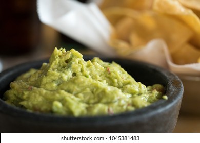 Macro close up on avocado guacamole in a black mortar bowl, with a basket of corn tortilla chips in the blurry background, at a Mexican restaurant