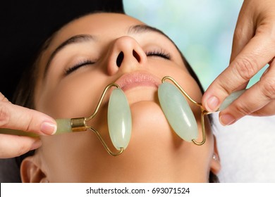 Macro close up portrait of woman having facial beauty treatment in spa. Therapist massaging chin with jade rollers.