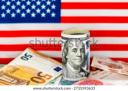 A macro close up of a roll of US dollar bills on top of other currency bank notes with the flag of United States of America in the background. A dollar hegemony concept.