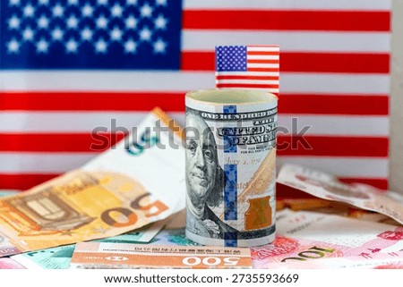 A macro close up of a roll of US dollar bills with a small flag of America placed on top of other currency bank notes, and the national flag of America in the background. A dollar hegemony concept.
