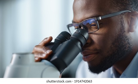 Macro Close Up Shot of a Handsome Black Male Scientist Wearing Glasses and Looking into the Microscope. Microbiologist Working on Molecule Samples in Modern Laboratory with Technological Equipment.