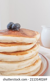 Macro Close Up Of Stack Of Pancakes With Blueberries On Top And Maple Syrup Or Honey Running Down The Sides. Pouring Jug In Background Set Against A Light Airy White Backdrop.