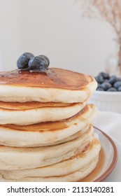 Macro Close Up Of Stack Of Pancakes With Blueberries On Top And Maple Syrup Or Honey Running Down The Sides. Blueberries And Flowers In Background Set Against A Light Airy White Backdrop.