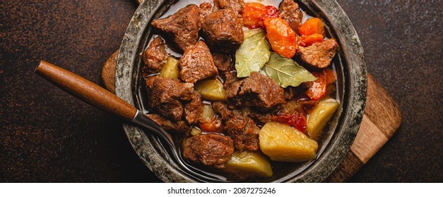 Macro Close-up Of Delicious Beef Meat Stew Dish With Meat Cubes, Potatoes, Carrot And Gravy In Rustic Metal Bowl On Brown Concrete Background Top View