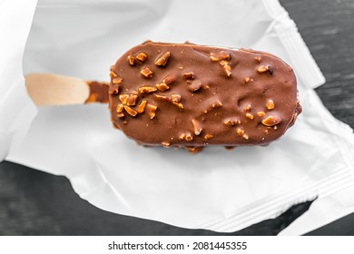 Macro closeup flat lay top view of one chocolate almond nuts ice cream bar on wood stick with plastic white wrap packaging package on kitchen table