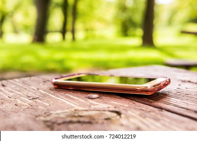 Macro Closeup Of Lost Generic Light Pink Rose Gold Smartphone, Phone Left, Forgotten In Plastic Clear Case Lying On Table In Outside Summer Park