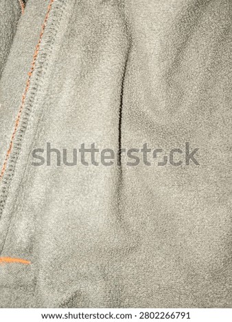 Macro close-up texture of soft dark grey polar fleece fabric featuring a contrasting orange zig-zag stitch seam on the edge, suitable for warm winter apparel backgrounds