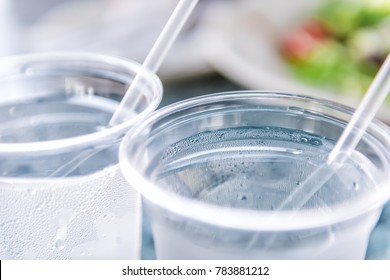 Macro Closeup Of Two Plastic Cups With Ice Cubes And Water And Condensation On Table With Straws By Green Salad In Restaurant