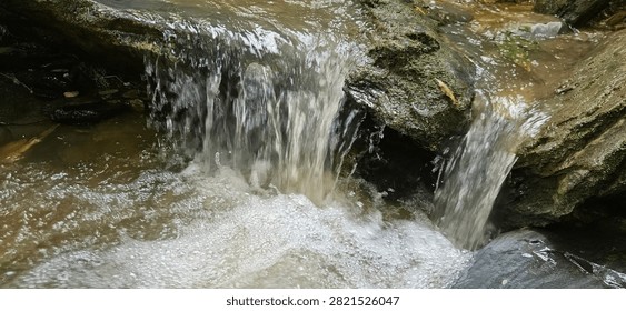 A macro close-up view of a small mountain waterfall cascading down a weathered rock step. The fast-flowing fresh water creates dense white foam and bubbles as it crashes into a turbulent natural pool below, highlighting the raw textures, fluid motion, and pristine energy of a wild stream hidden within the jungle