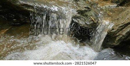 A macro close-up view of a small mountain waterfall cascading down a weathered rock step. The fast-flowing fresh water creates dense white foam and bubbles as it crashes into a turbulent natural pool below, highlighting the raw textures, fluid motion, and pristine energy of a wild stream hidden within the jungle
