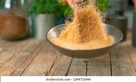 MACRO, DOF: Heap of stale bread crumbs getting toasted in a metal pan. Unrecognizable chef roasting grated bread and crackers above a wooden table. Golden breadcrumbs falling onto the kitchen counter.