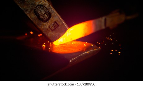 MACRO, DOF: Small black particles flying away from hot red blade while getting forged by an unrecognizable blacksmith. Glowing piece of iron being forged into blade. Person forging knife blades.