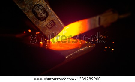 MACRO, DOF: Small black particles flying away from hot red blade while getting forged by an unrecognizable blacksmith. Glowing piece of iron being forged into blade. Person forging knife blades.