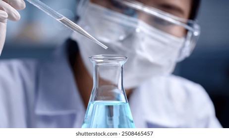 Macro front angle view of a scientist is dropping light blue liquid substance into an erlenmeyer flask. Concept of substance study experiment on blurred lab background, for promotional photo