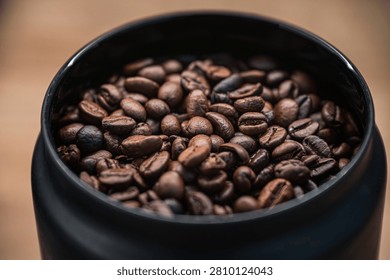 A macro, high-angle shot looking into a sleek black storage canister filled to the brim with aromatic medium-dark roasted coffee beans. The image showcases rich textures and details of the coffee grains against a soft, out-of-focus background.