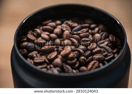 A macro, high-angle shot looking into a sleek black storage canister filled to the brim with aromatic medium-dark roasted coffee beans. The image showcases rich textures and details of the coffee grains against a soft, out-of-focus background.