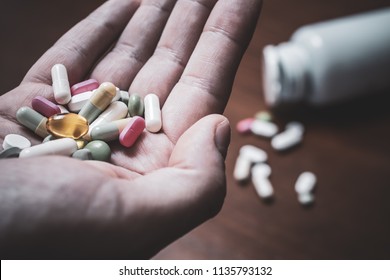 Macro Of A Male Hand Holding Several Pills With Prescription Bottle And More Pills In Background - Taking Too Many Pills Concept