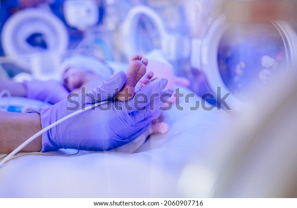Macro photo of doctor\'s hands and legs of a child.\
Newborn is placed in a medical incubator under ultraviolet lamp.\
Neonatal intensive care\
unit.